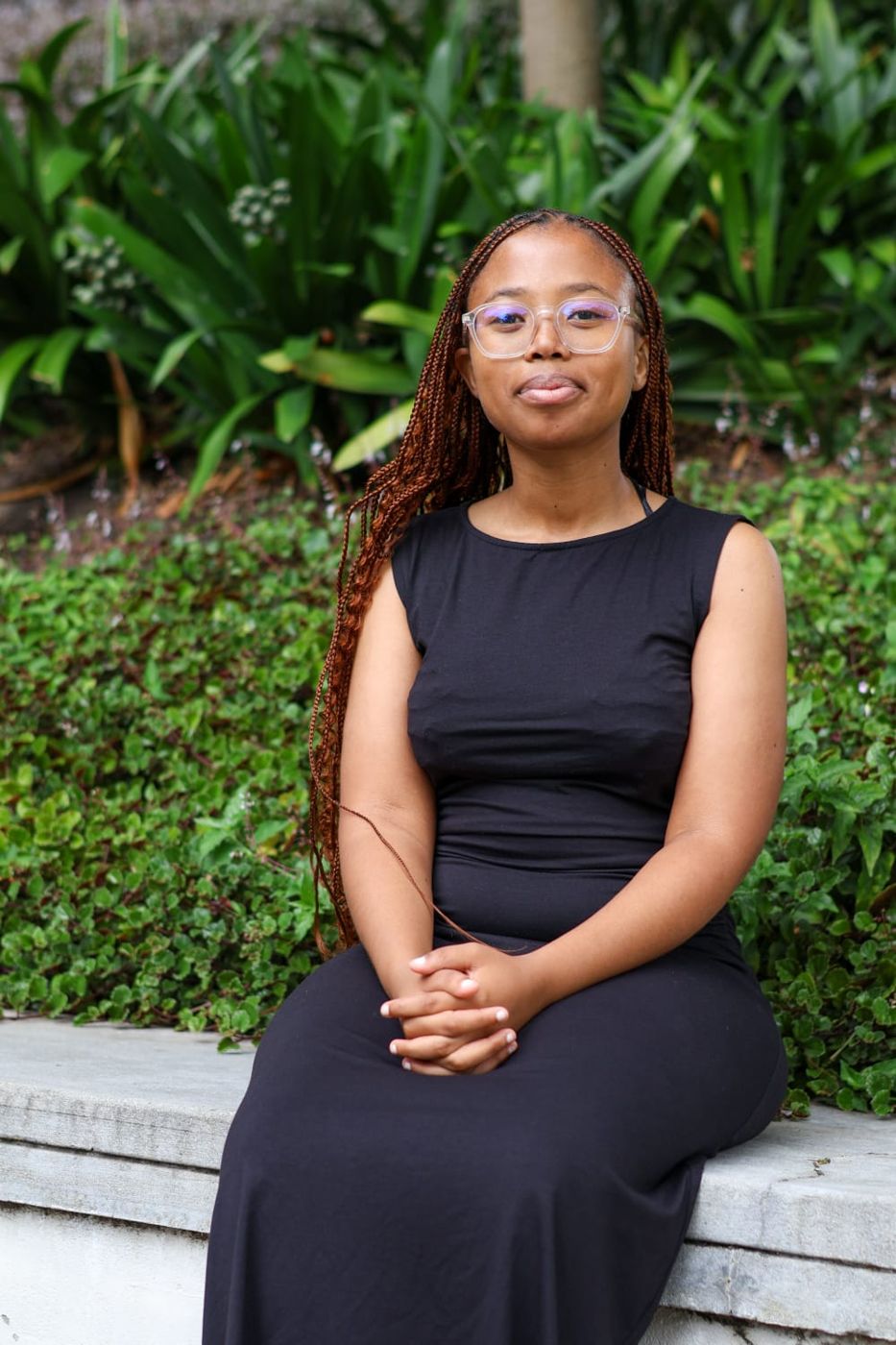 Okuhle Ncinane sitting on a low wall in front of a green garden, smiling softly